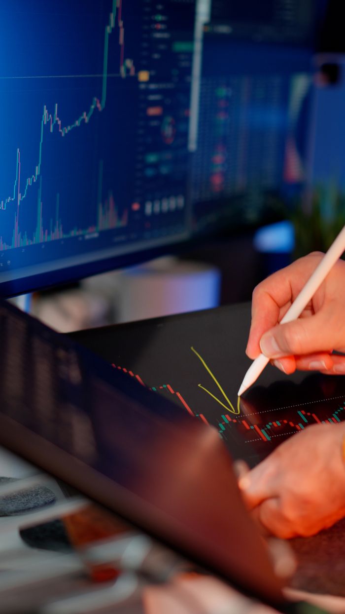 about-01 Person using a stylus on a tablet for stock market analysis, with charts on a monitor.