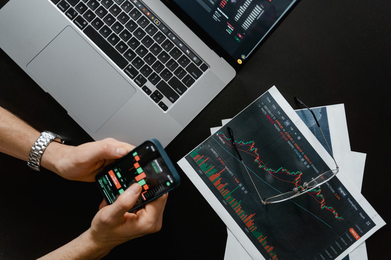 hero-01 Hands using smartphone beside laptop with stock charts, showcasing digital trading.