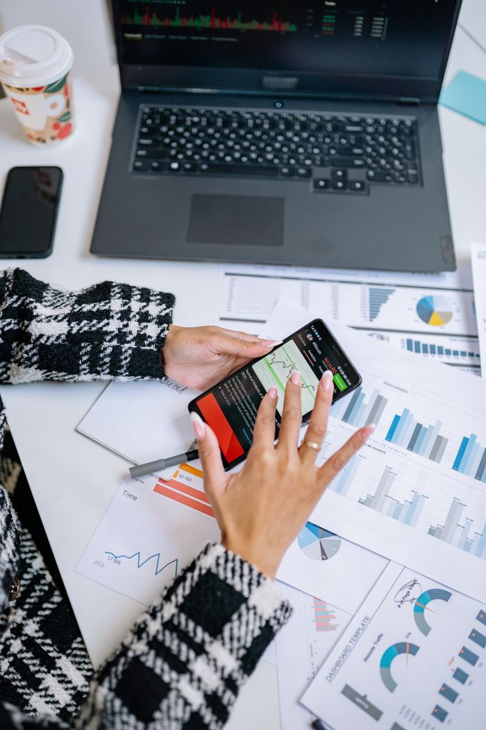 why-choose-us-03 Person analyzing business charts with smartphone and laptop on desk.